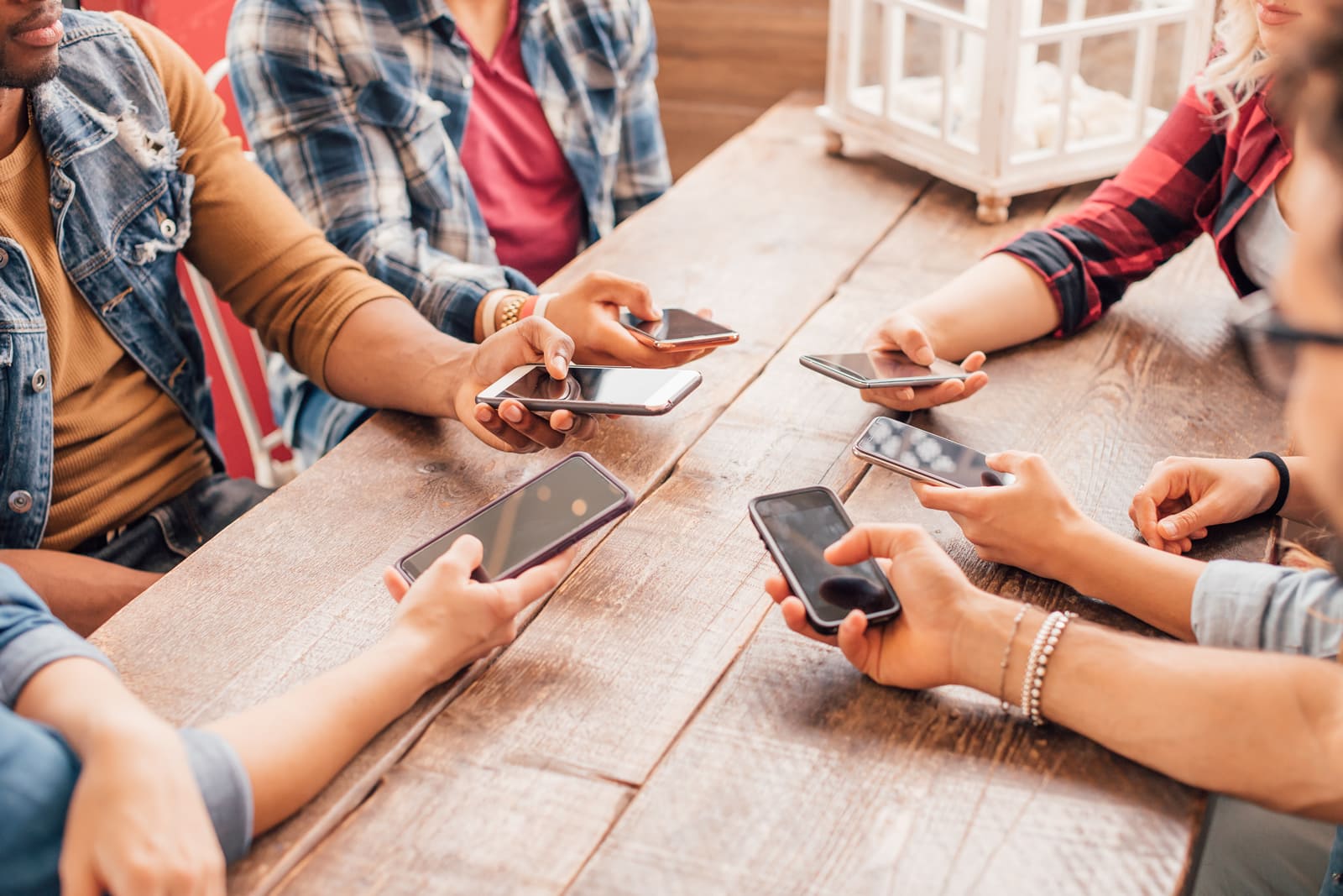 Group of young friends participating in an online bulletin board on their smartphones while sitting at a wooden table